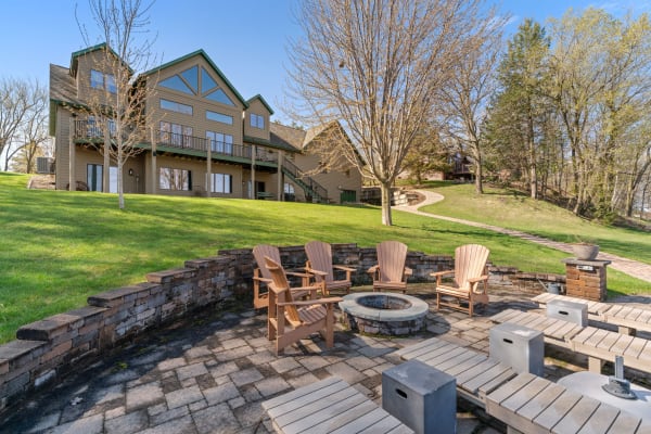 An outdoor seating area with a fire pit, Adirondack chairs, and a large house in the background surrounded by greenery.