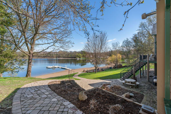 Lakeside scene with a paved path, sandy shore, and a dock under a clear blue sky.