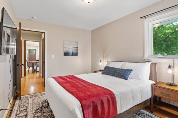 A cozy bedroom featuring a queen-sized bed with a red throw blanket, wooden nightstands, and a window with views of green trees.