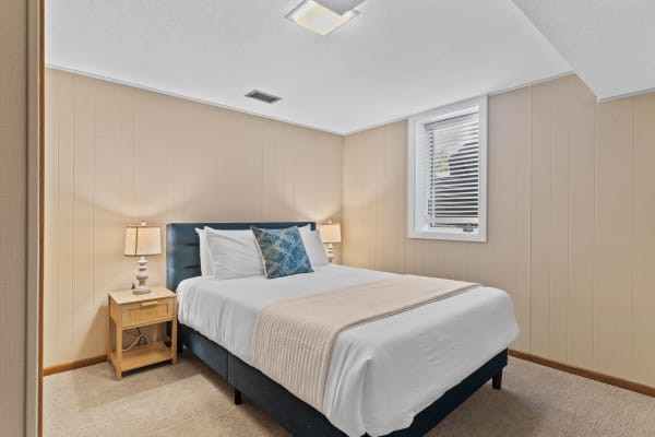 A small bedroom with a queen bed, bedside lamp, and sunlight coming through the window.