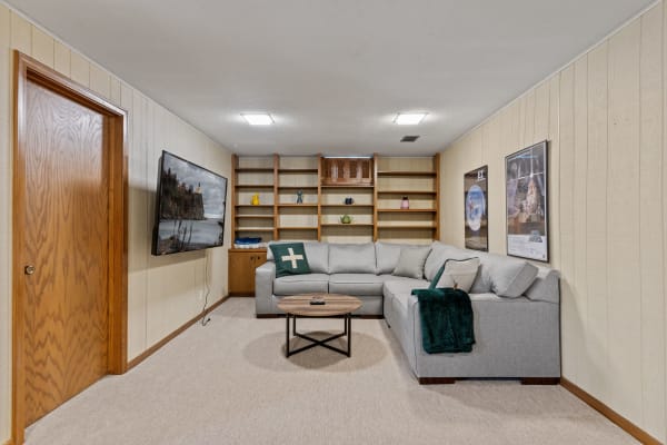 A well-furnished basement with a gray couch, round table, TV, and decorative shelves.