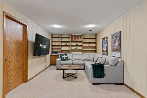A comfortable basement with a gray sectional sofa, wooden shelves, and movie posters on the wall.