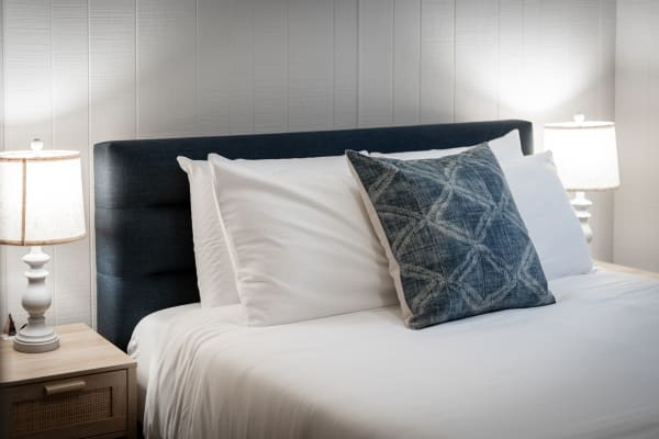 A bed with a dark headboard, white pillows, and geometric blue decorative pillow, accompanied by lamps on wooden nightstands.