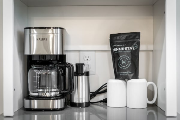 A modern coffee station with KRUPS coffee machine, frother, MINNESTAY coffee bag, and white mugs.