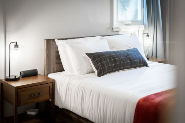 A neatly made bed with white linens and a plaid pillow, accompanied by a lamp and a speaker on a nightstand.