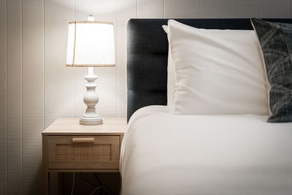 A bedside table with a lamp beside a well-made bed with white linens and a dark headboard.