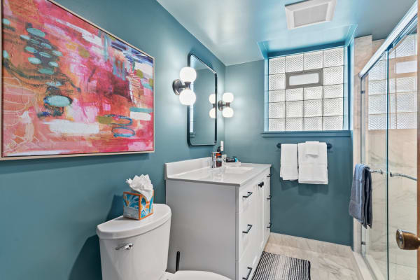 A modern bathroom with teal walls, a white vanity, abstract artwork, and a glass shower.