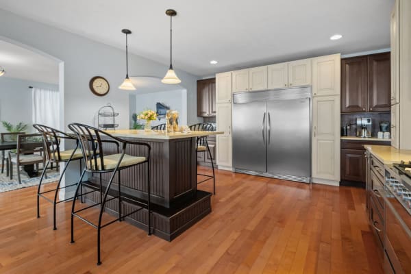A warm kitchen with an island, bar stools, stainless steel refrigerator, and wooden floors.