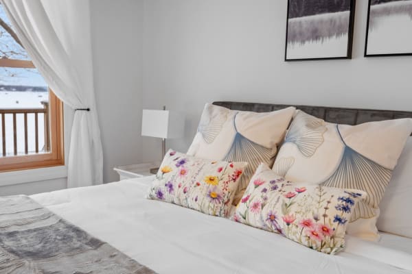 A beautifully decorated bedroom with floral accent pillows, a grey headboard, and a snowy landscape visible through a window.