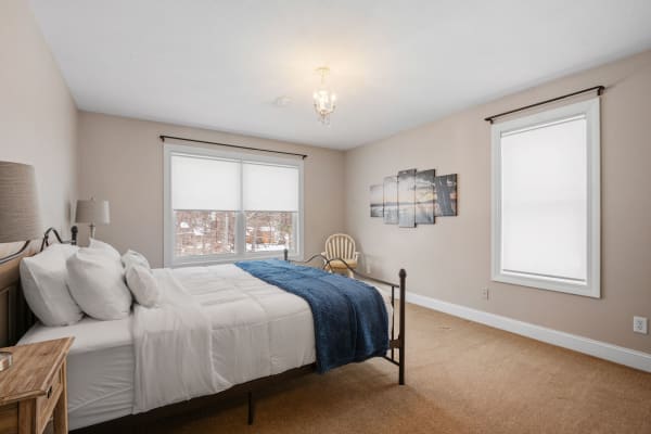 A tranquil bedroom featuring a dark metal bed, soft white bedding with a blue blanket, a wooden nightstand with a lamp, large windows, and wall artwork.