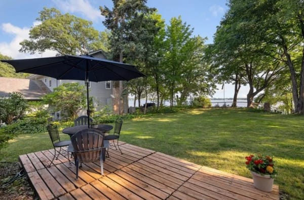 Outdoor wooden deck with table and chairs, surrounded by greenery and a view of the water.