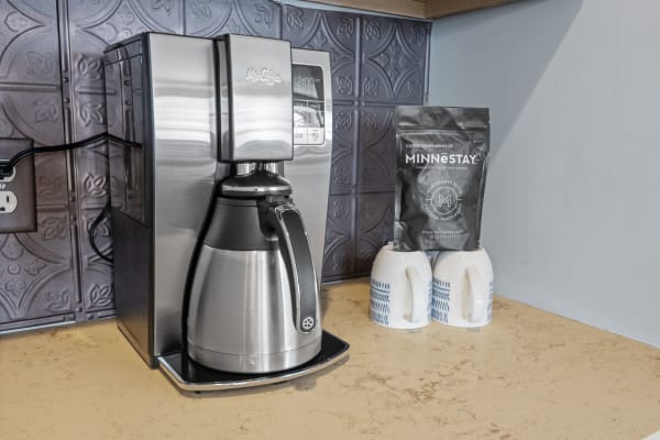 A stainless steel coffee maker with a thermal carafe, a coffee bag, and two patterned mugs on a countertop.