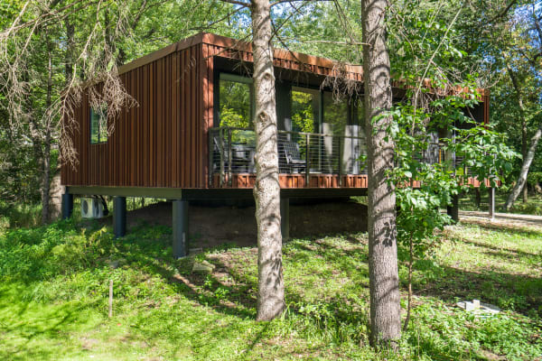 Modern elevated house with a rust-colored exterior and large windows, surrounded by trees and greenery.