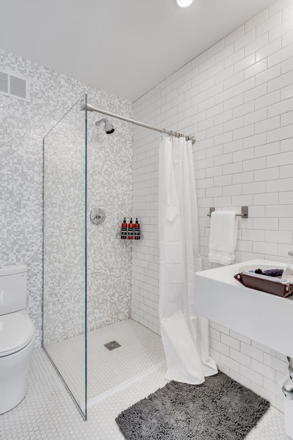 A contemporary bathroom with a glass shower, white tiles, and a dark gray bath mat.