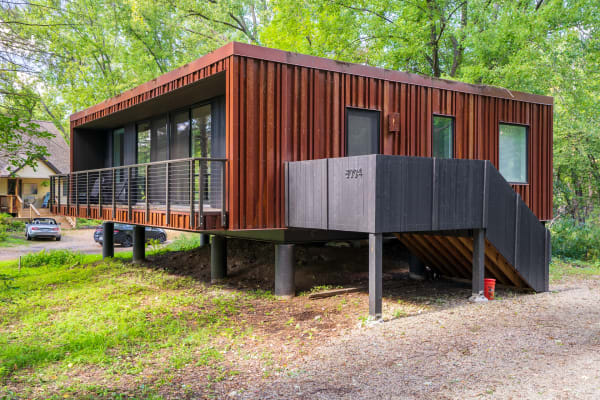 An elevated modern house with rust-colored siding and large glass windows surrounded by greenery.