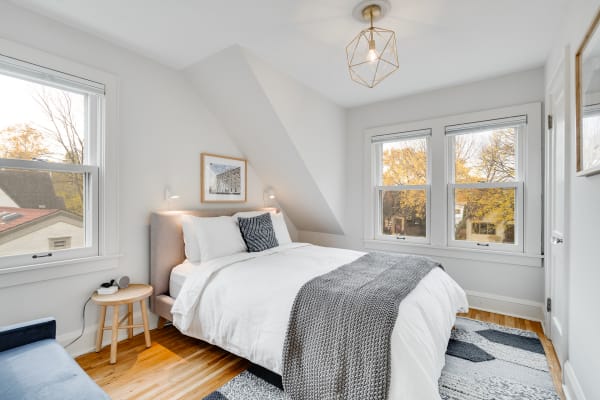 A cozy bedroom with a bed, a geometric light fixture, and autumn trees visible through the windows.