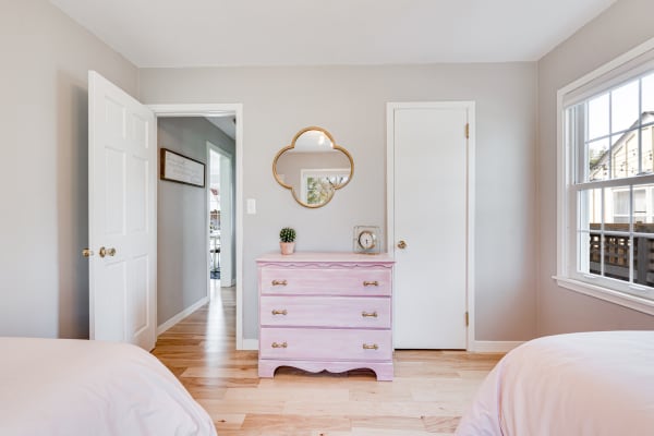 A light-filled bedroom with a pink dresser, gray walls, and wooden floors.