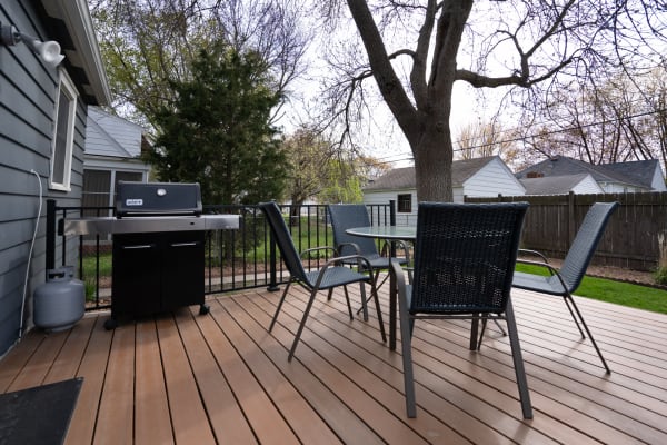 An outdoor deck with a grill, a round table, and four chairs surrounded by greenery.