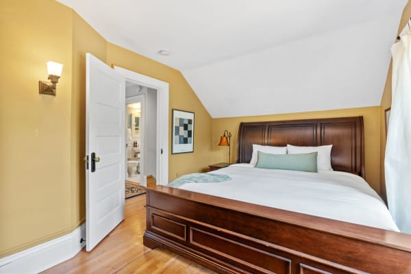 A cozy bedroom with a wooden bed and golden-yellow walls, showing an adjoining bathroom in the background.