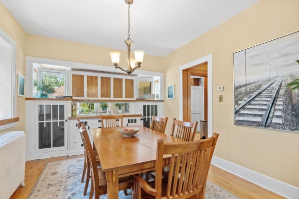A cozy dining room featuring a wooden table with chairs, a fruit bowl, and large windows connecting to a kitchen.