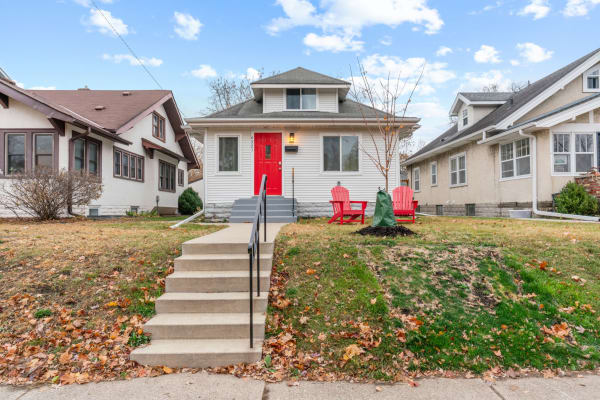 A white bungalow with a red door and two red chairs on the porch.