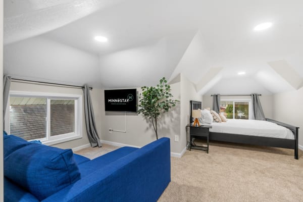 A modern attic bedroom featuring a blue couch, a bed with white linens, a potted plant, and a mounted TV.
