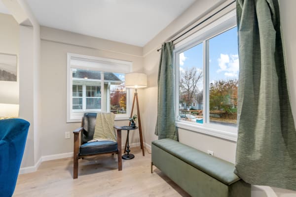 A corner of a room with a black armchair, side table, floor lamp, and a view of colorful trees outside the window
