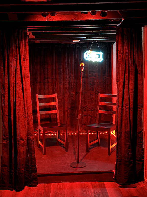 A small stage setting featuring two chairs, a microphone, and red curtains with warm lighting.