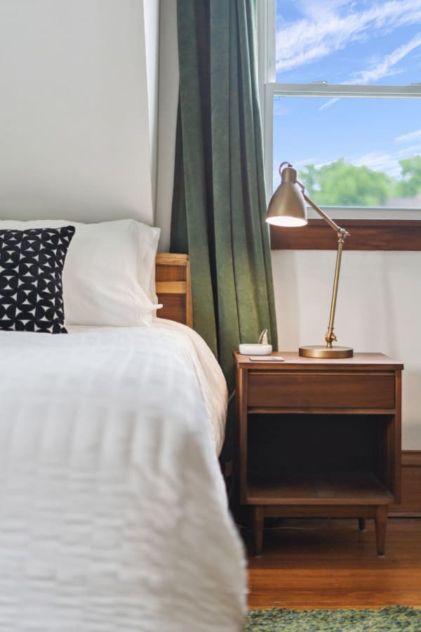A warm bedroom corner with a bed, a patterned pillow, a nightstand, and a window with green curtains.