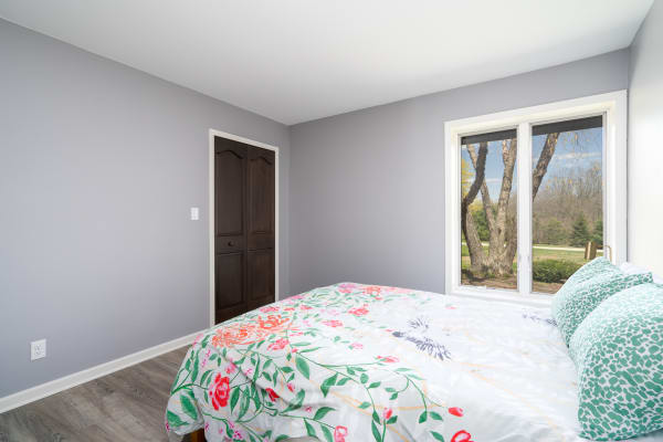 A bedroom featuring gray walls, a floral duvet on the bed, and a large window with a view of trees.