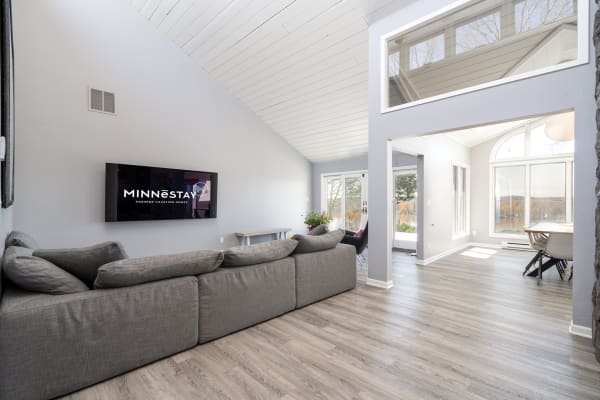 A spacious, contemporary living room with a gray sofa, a TV on the wall, and large windows letting in natural light.