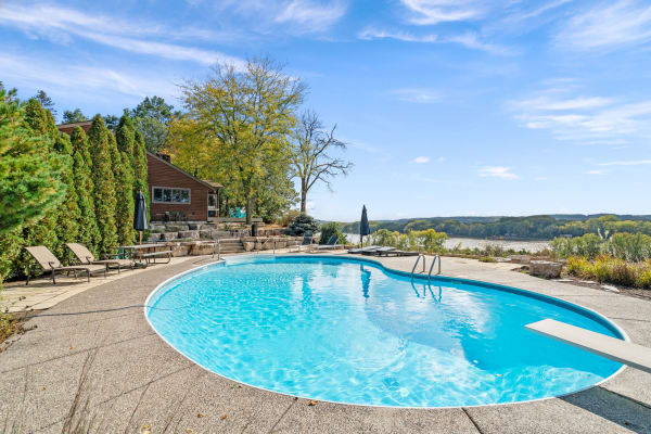 Outdoor swimming pool with clear blue water, surrounded by patio and trees, overlooking a river and hills.