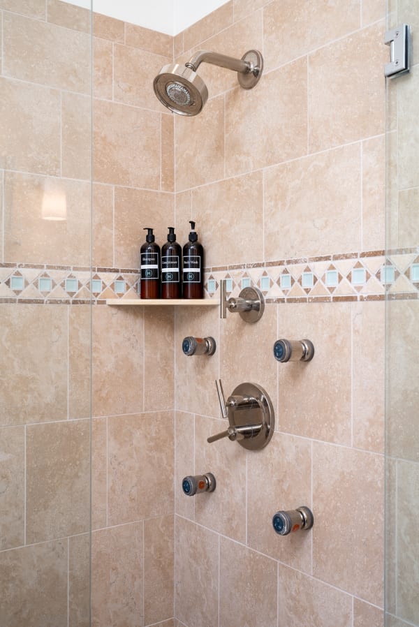 A minimalist shower with stone tiles, a chrome showerhead, and a wooden shelf holding shampoo and body wash.