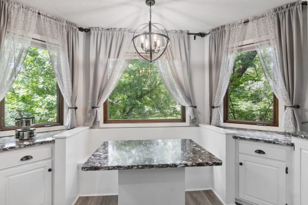 A kitchen nook with large windows and gray curtains, featuring a marble-patterned table and a modern chandelier.