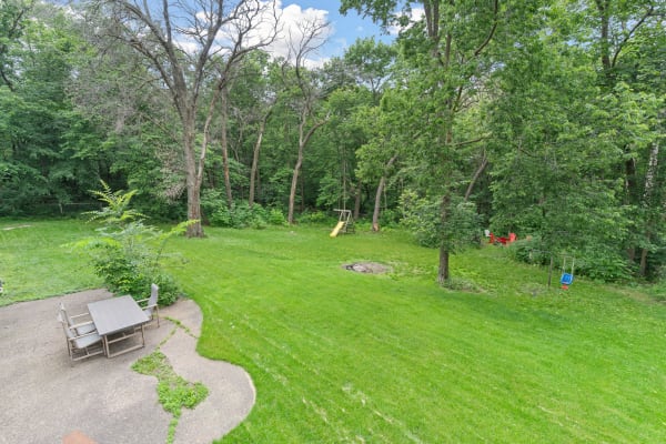 A backyard featuring green grass, a patio table, a playground, and dense trees.