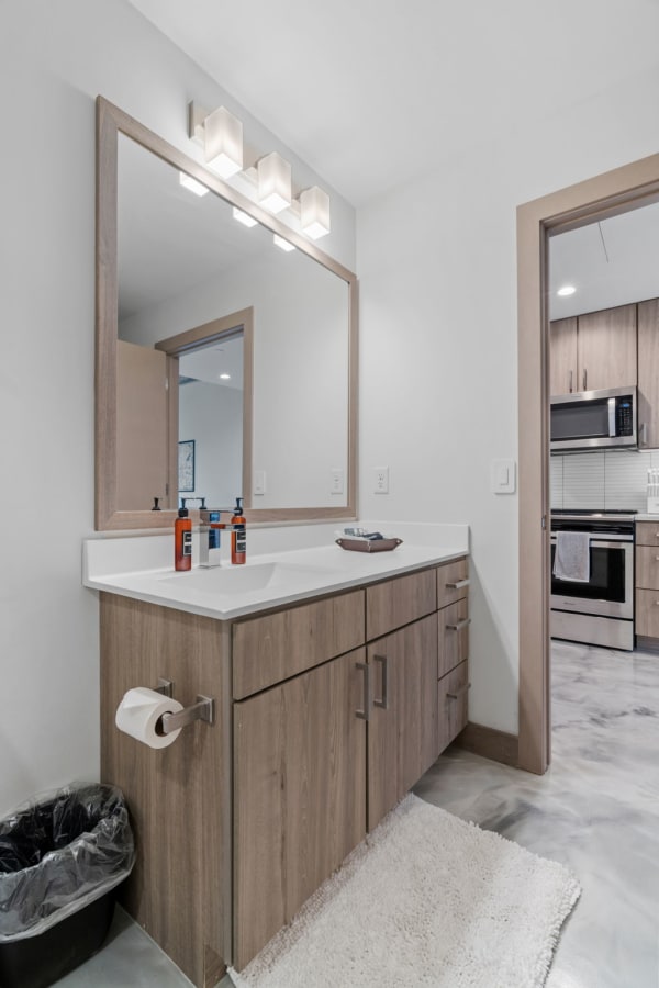 A modern bathroom vanity with a large mirror, light fixtures above, and a glimpse of a kitchen in the background.