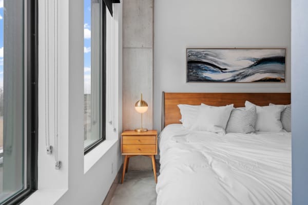 A bright, modern bedroom showing a bed with white linens, stylish lamp, and abstract art above.