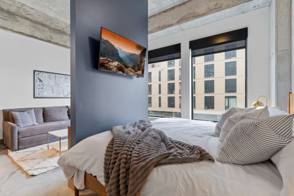 A modern studio bedroom featuring a bed with gray blanket, a sofa, and a large window with an urban view.