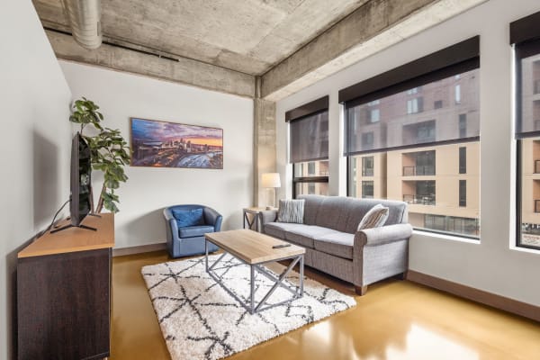 A contemporary living room with a gray sofa, blue armchair, and a wooden coffee table, featuring large windows and a cityscape painting.