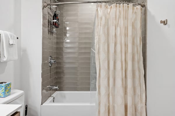 A modern bathroom with a bathtub and shower, light grey tiled walls, and cream shower curtain.