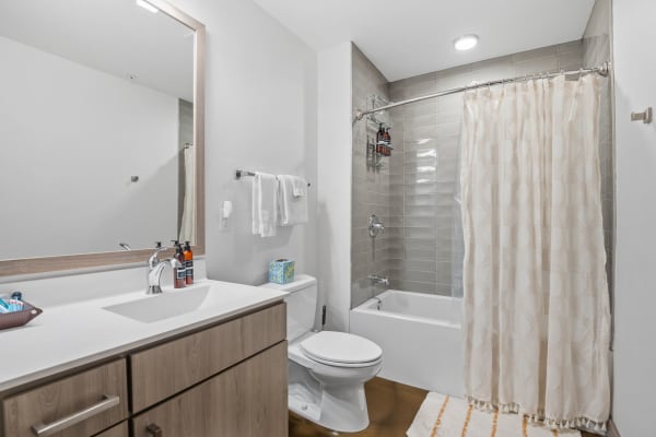 A spacious bathroom featuring a white toilet, light wood vanity, gray shower tiles, and a decorative shower curtain.