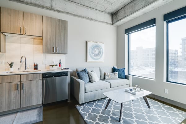 A stylish modern studio apartment with a gray sofa, kitchen area, and large windows.