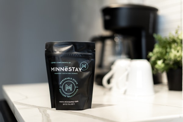 A black coffee pouch from MINNESTAY on a marble kitchen countertop with a coffee maker and white mugs in the background.