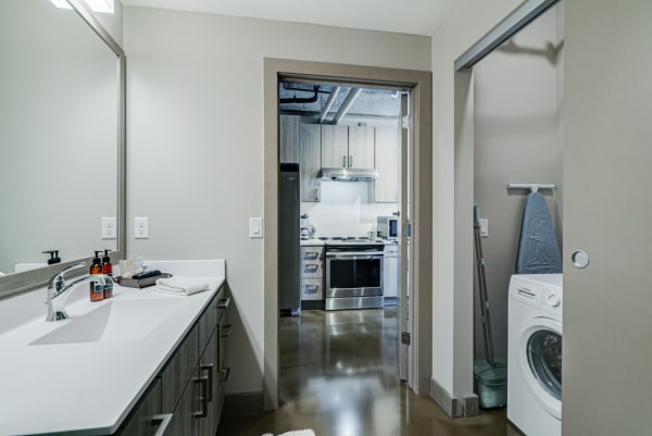 View of a modern bathroom with a laundry nook, showcasing a white vanity and stainless steel kitchen in the background.