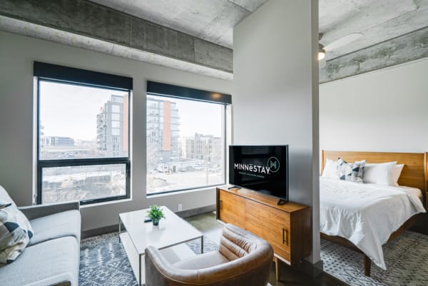 Modern studio apartment with a sofa, coffee table, television, and a bed, featuring large windows with an urban view.