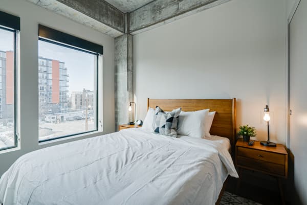 A modern bedroom with a wooden bed, white bedding, large windows, and an exposed concrete ceiling.