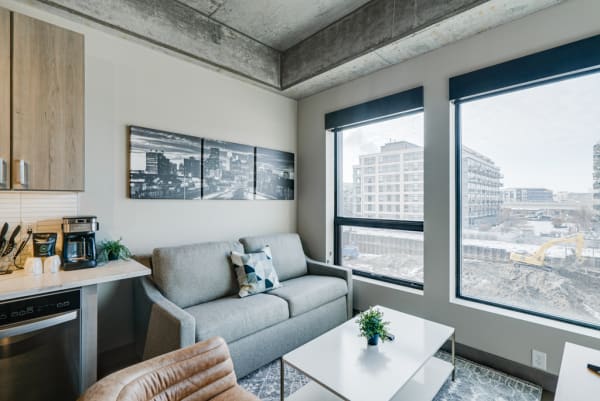 Modern living room with a gray sofa, kitchenette, and large windows overlooking a construction site.