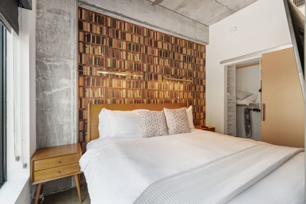 A modern bedroom featuring a bookshelf wallpaper, a neatly made bed, and a wooden nightstand.