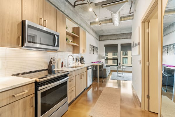 A contemporary kitchen featuring wood cabinets and stainless steel appliances, leading to a cozy living space with a sofa and table.