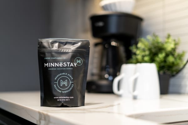 Black coffee bag labeled Minnestay on a stylish countertop next to coffee mugs and a plant.